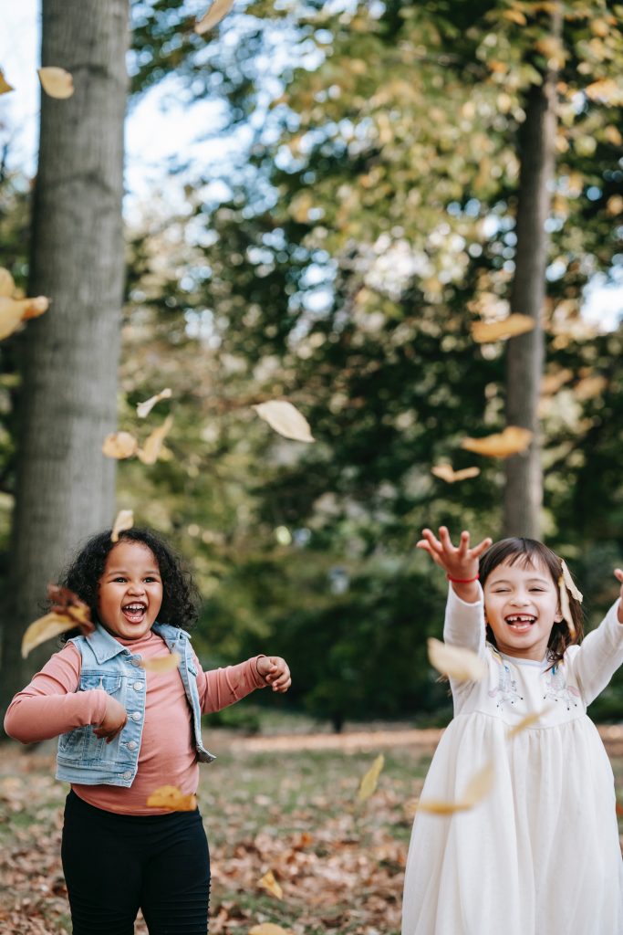 Two girls playing in woods with falling leaves. Photo by Charles Parker via Pexels